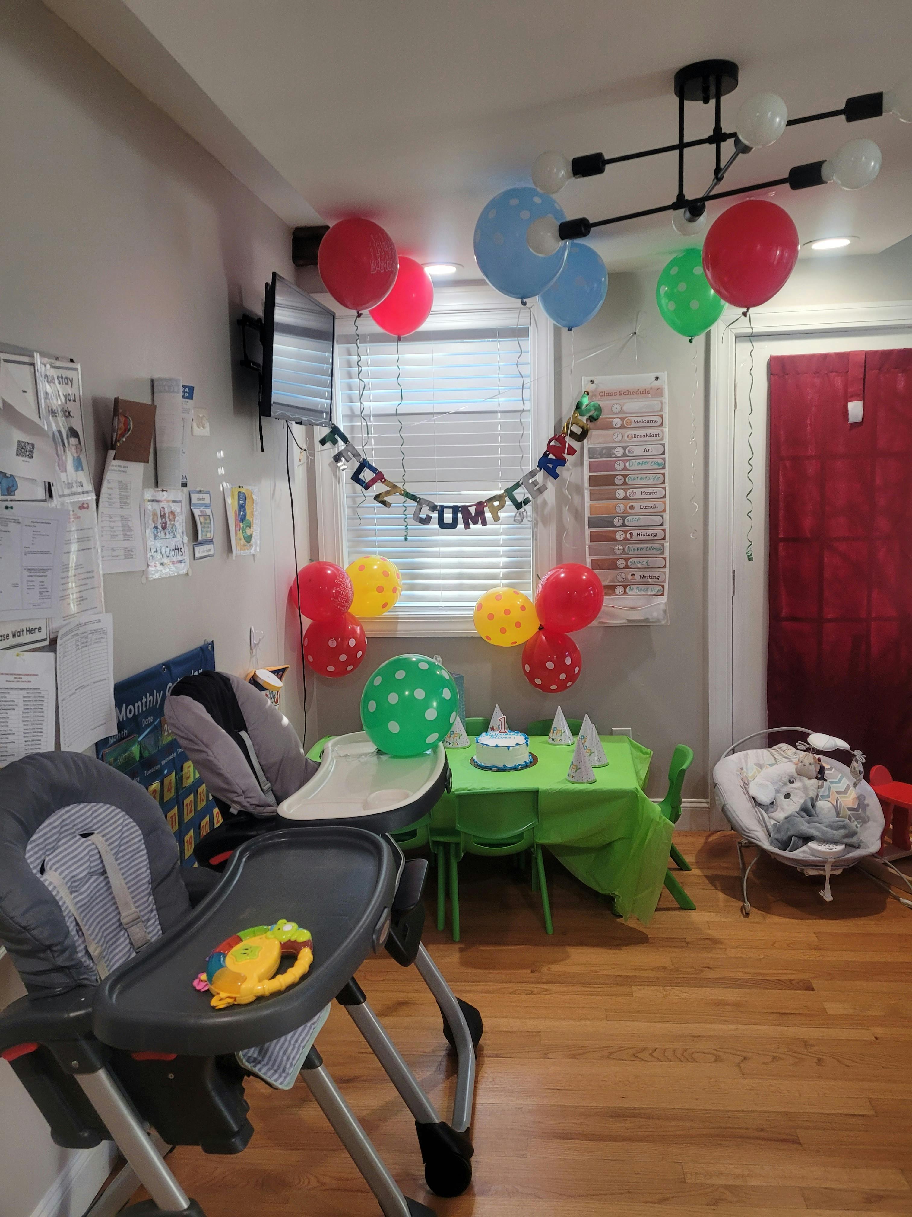 Balloons and a 'Feliz Cumpleaños' banner decorating a room with a high chair and baby bouncer