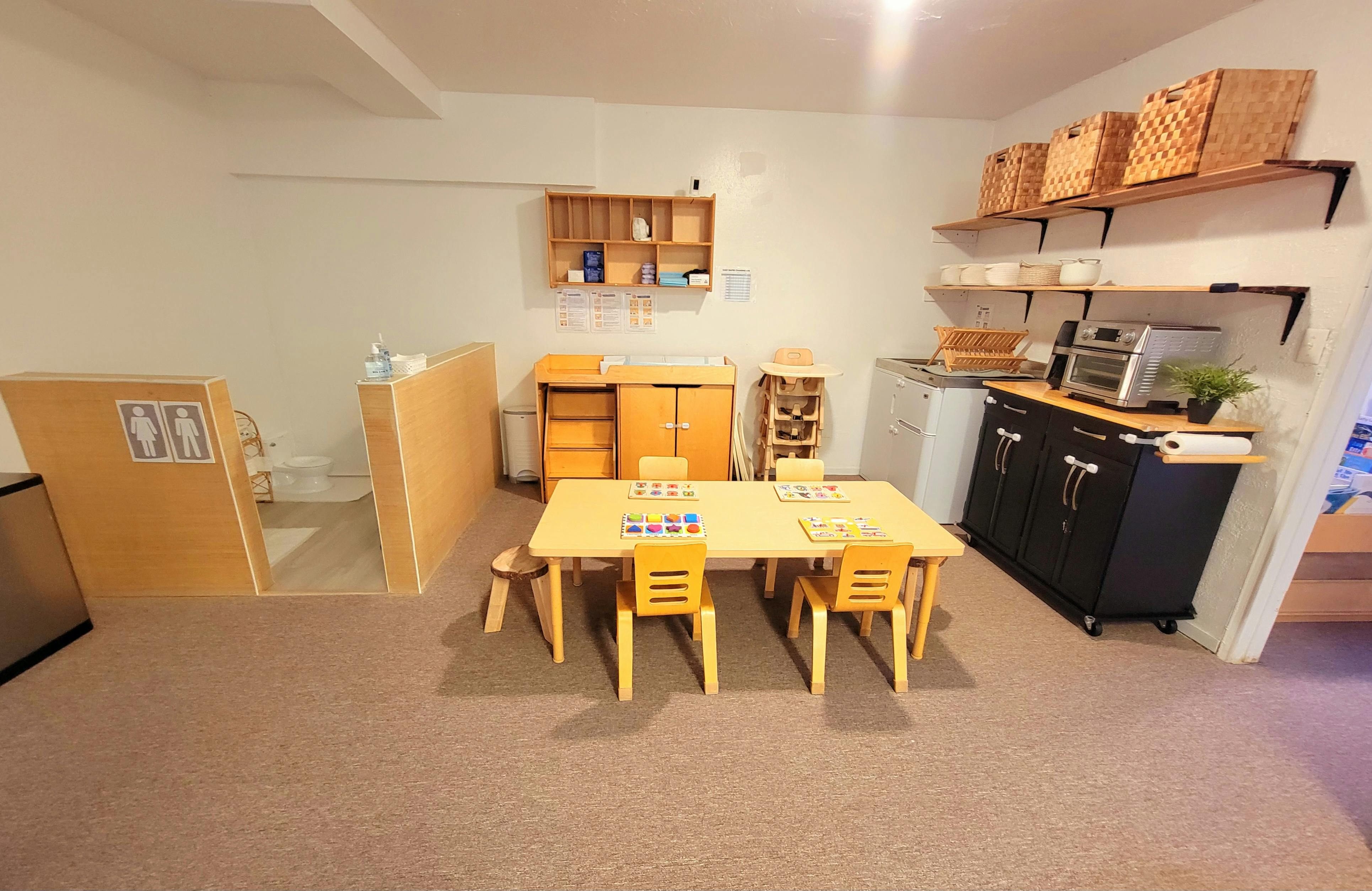 Classroom with small wooden tables and chairs, shelves, and a kitchen area