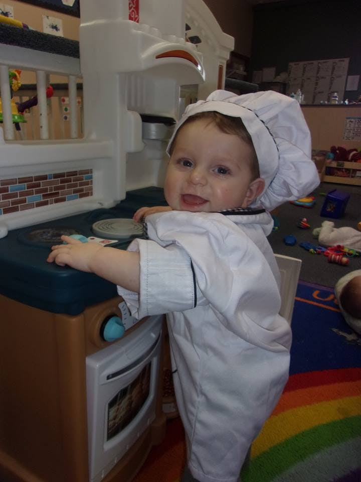 Child in chef's hat and coat playing with a toy kitchen
