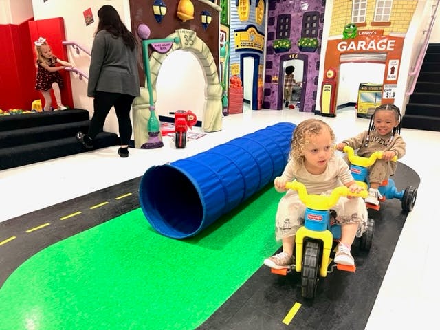 Two children riding tricycles on a play road with a tunnel