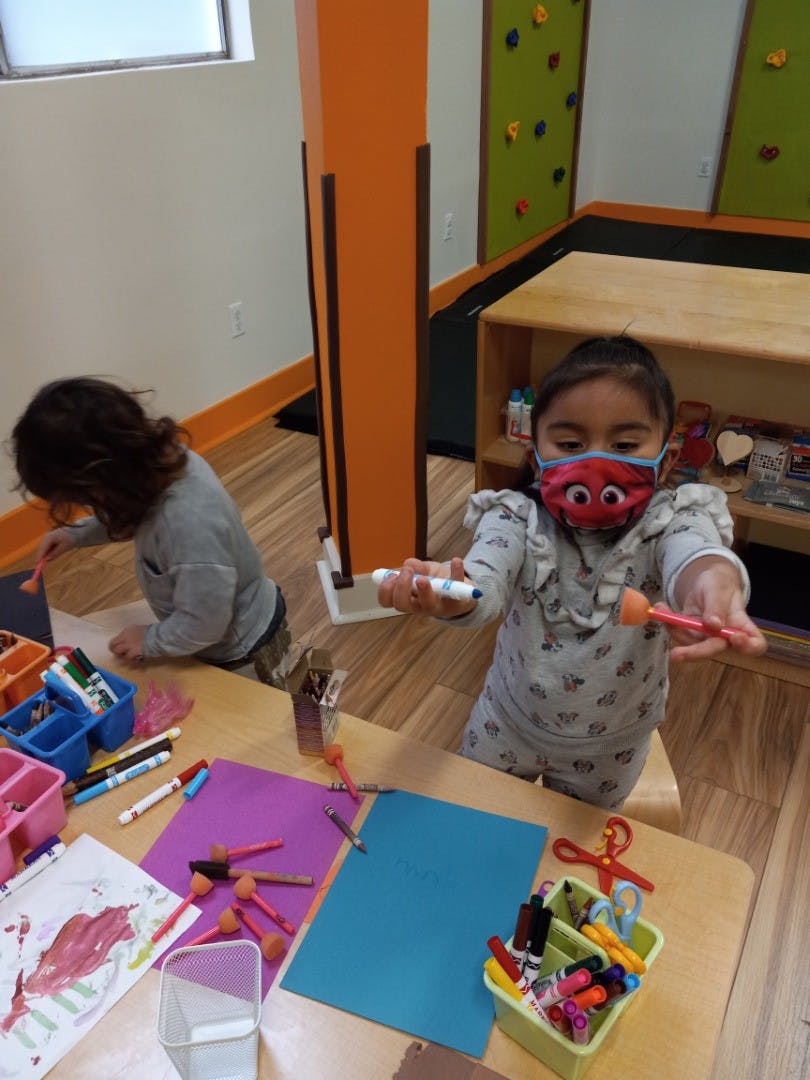 Child in a mask holding a marker and a paint brush, another child drawing