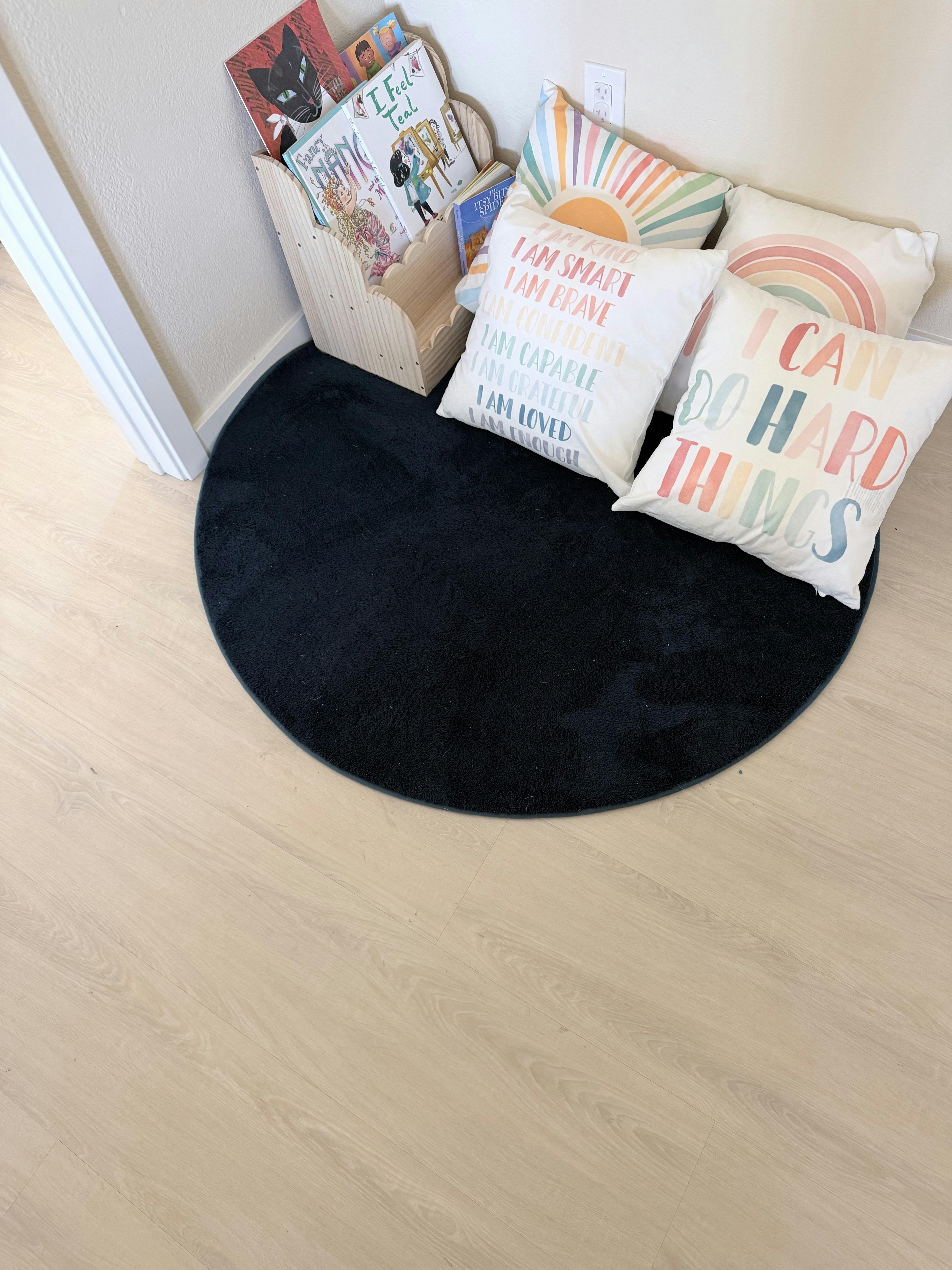 A reading corner with a round black rug, book rack, and inspirational pillows
