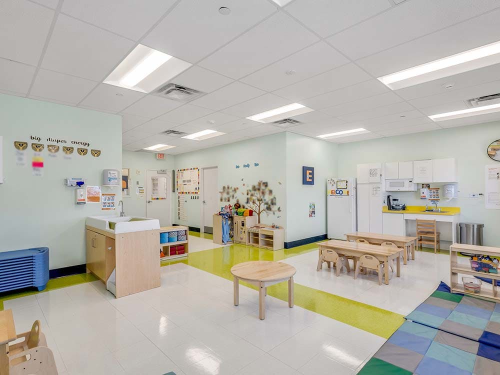 A brightly lit classroom with small wooden tables and chairs, a changing station, and a kitchen area