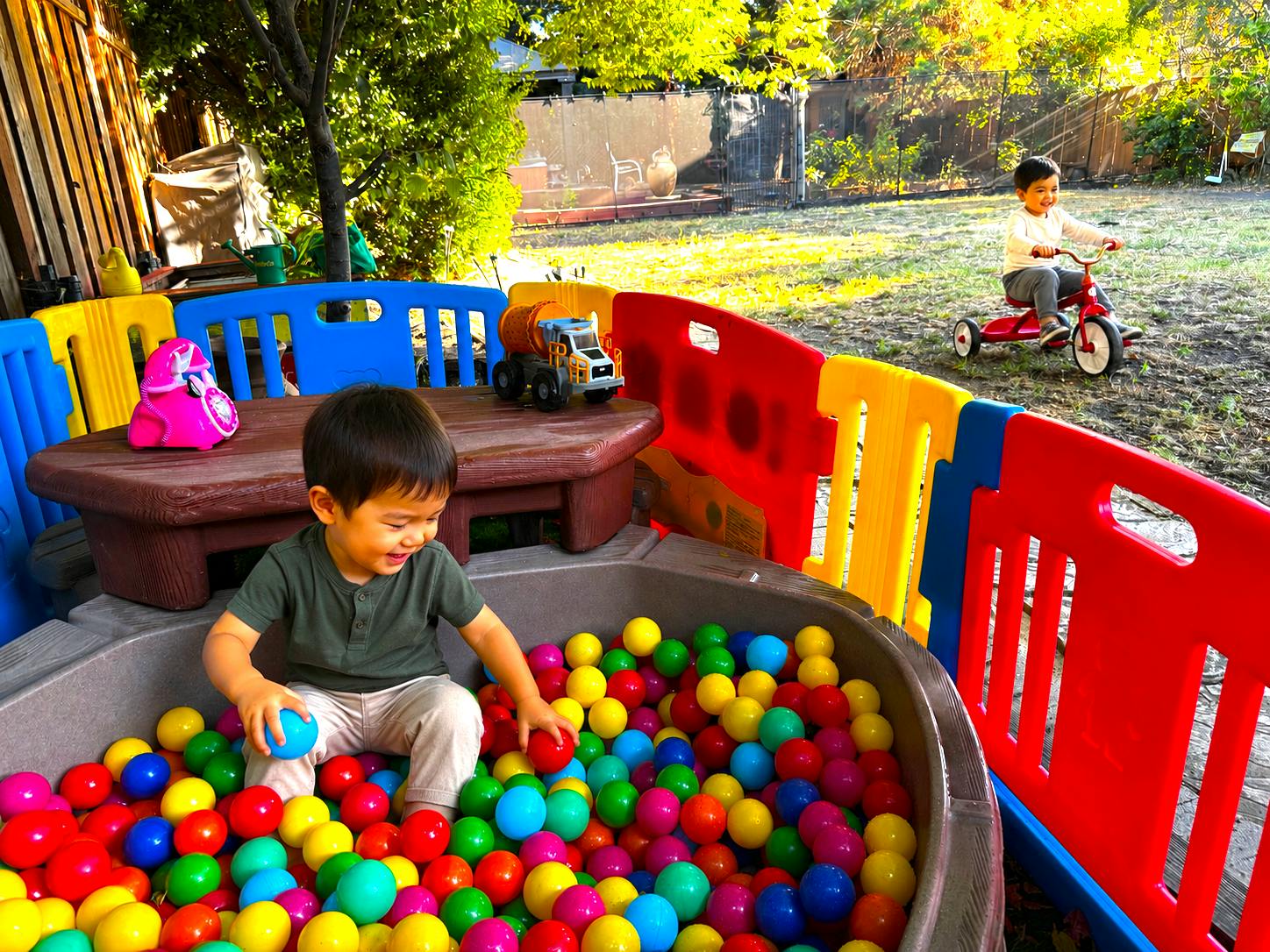 Child in a ball pit and another child riding a tricycle in a yard