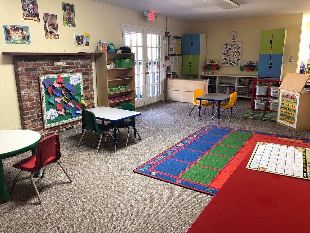 Empty classroom with small tables and chairs, colorful rug, and brick fireplace