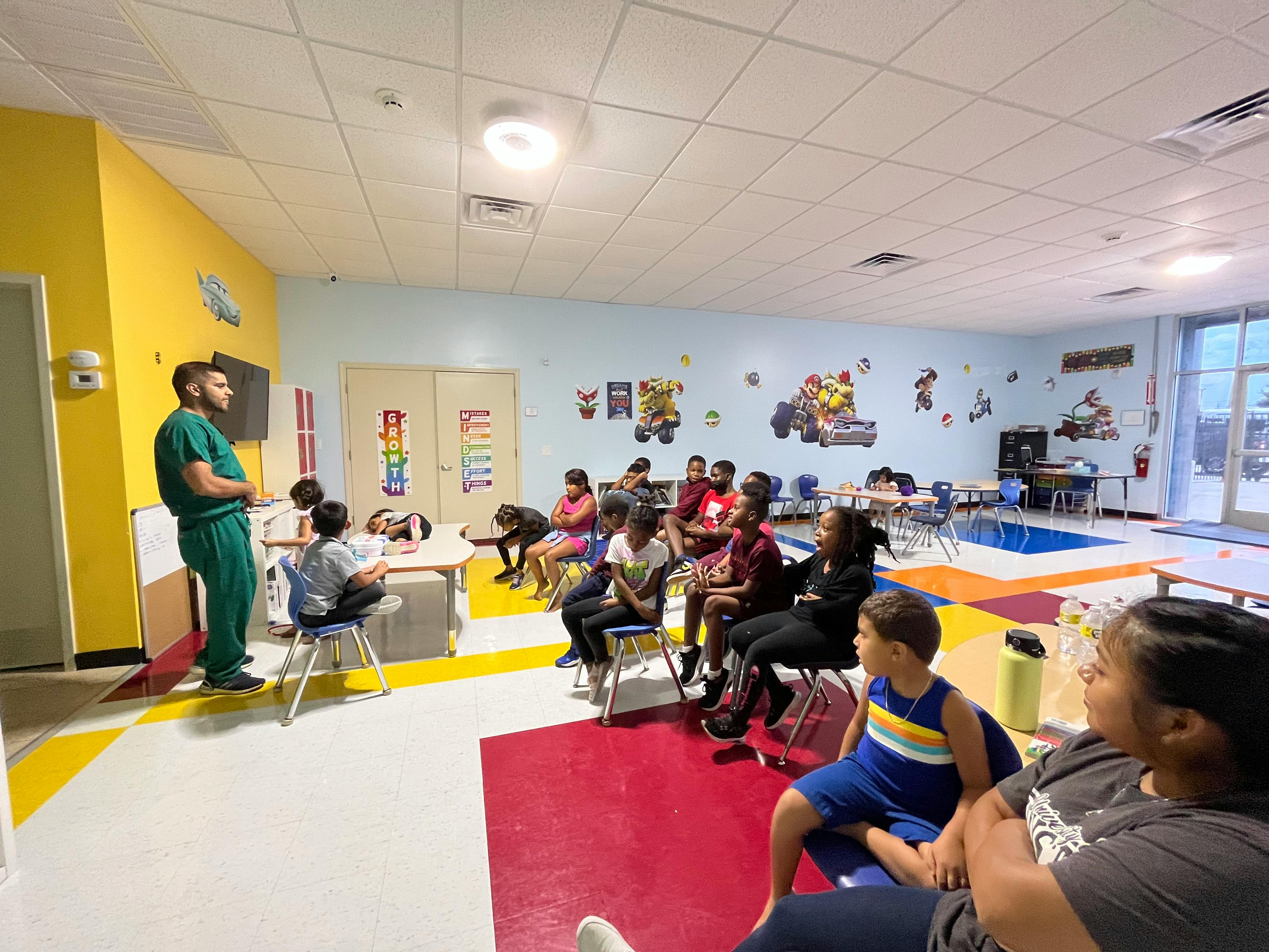 Person in scrubs speaking to a group of children and adults in a room