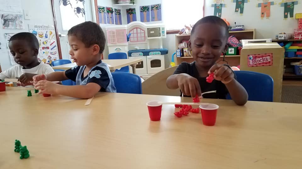 Three children playing with small objects at a table in a classroom