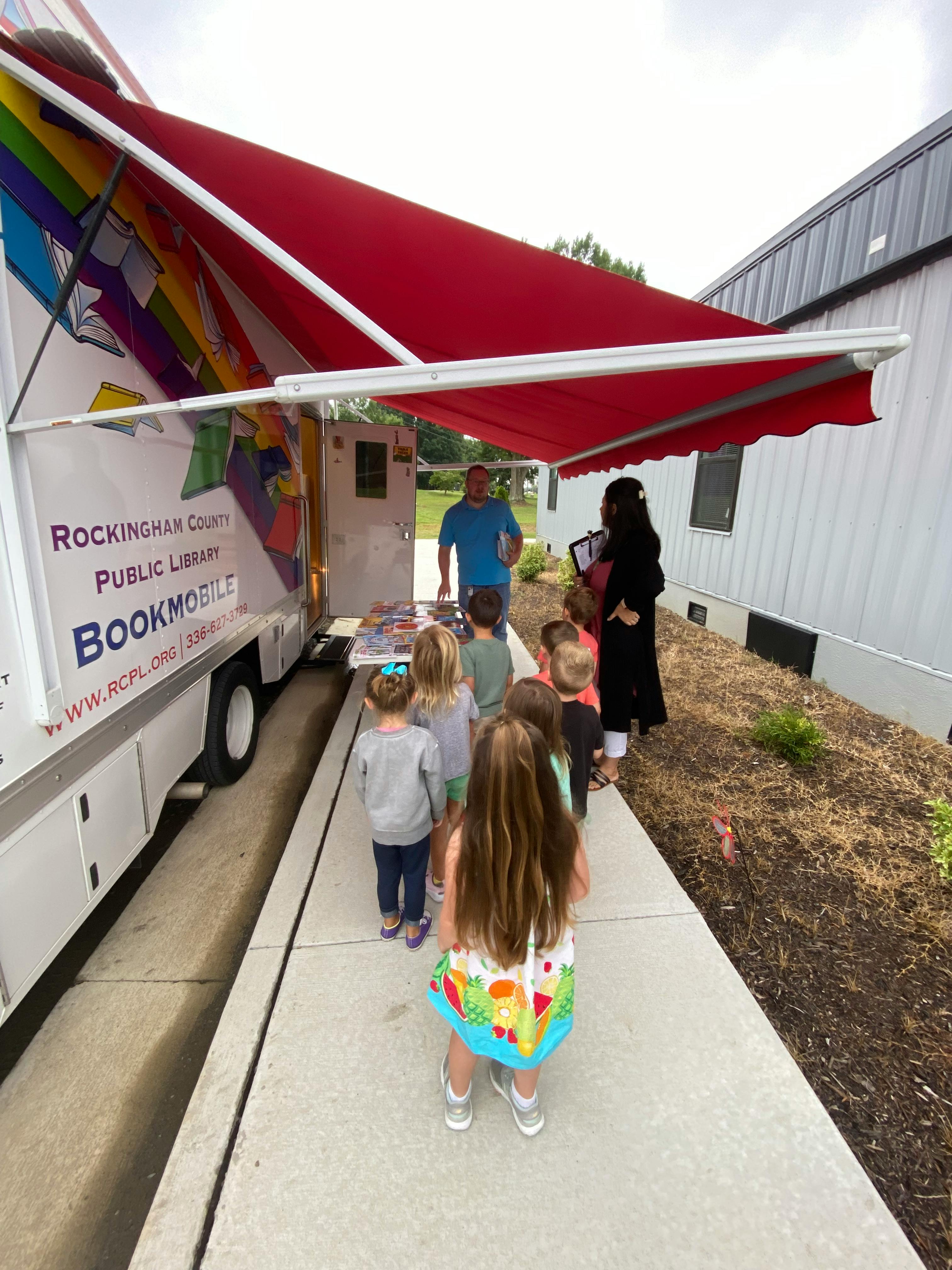 Children lining up to enter a bookmobile with a red awning
