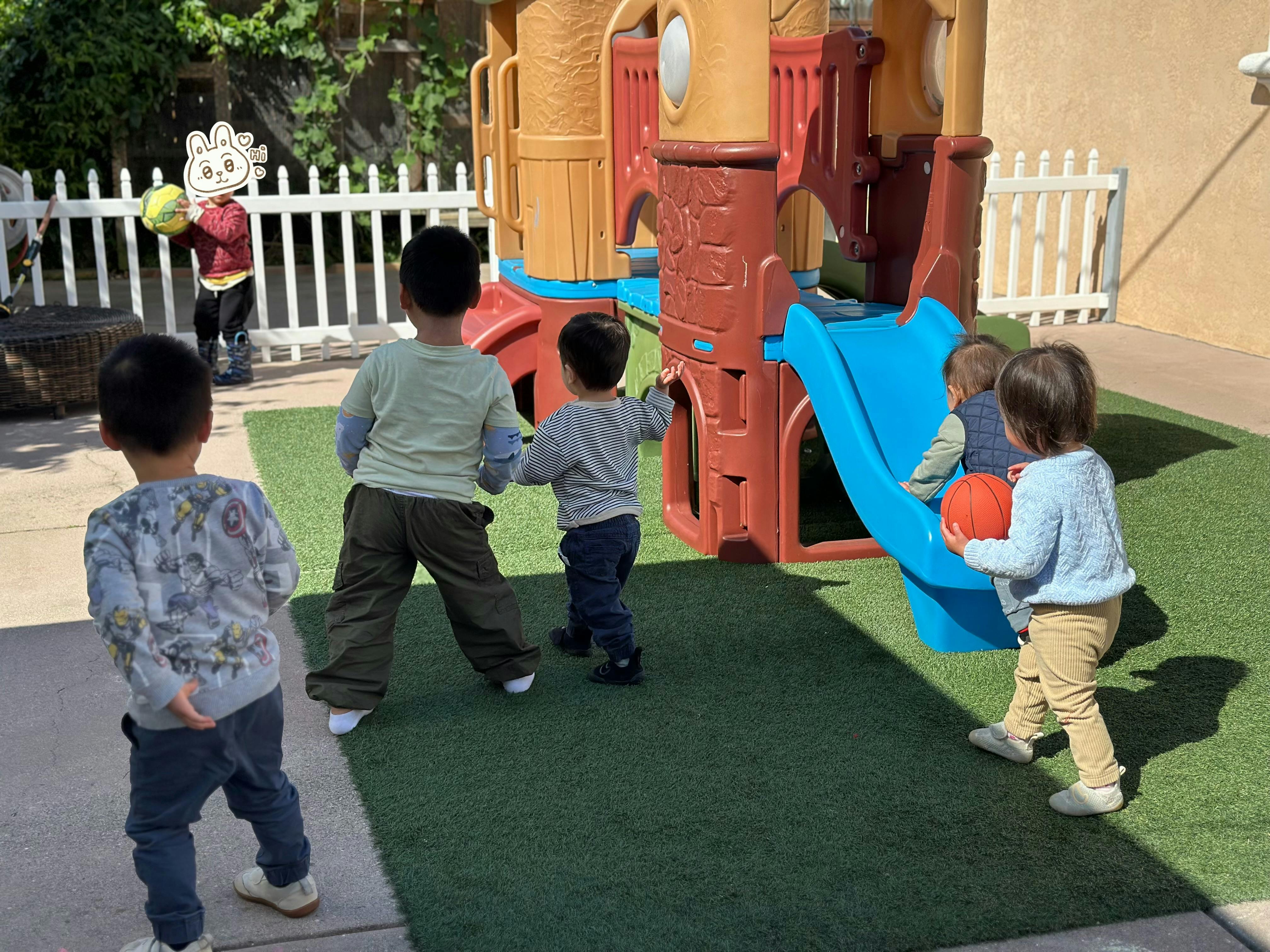 Several young children playing on green turf near a plastic playground structure
