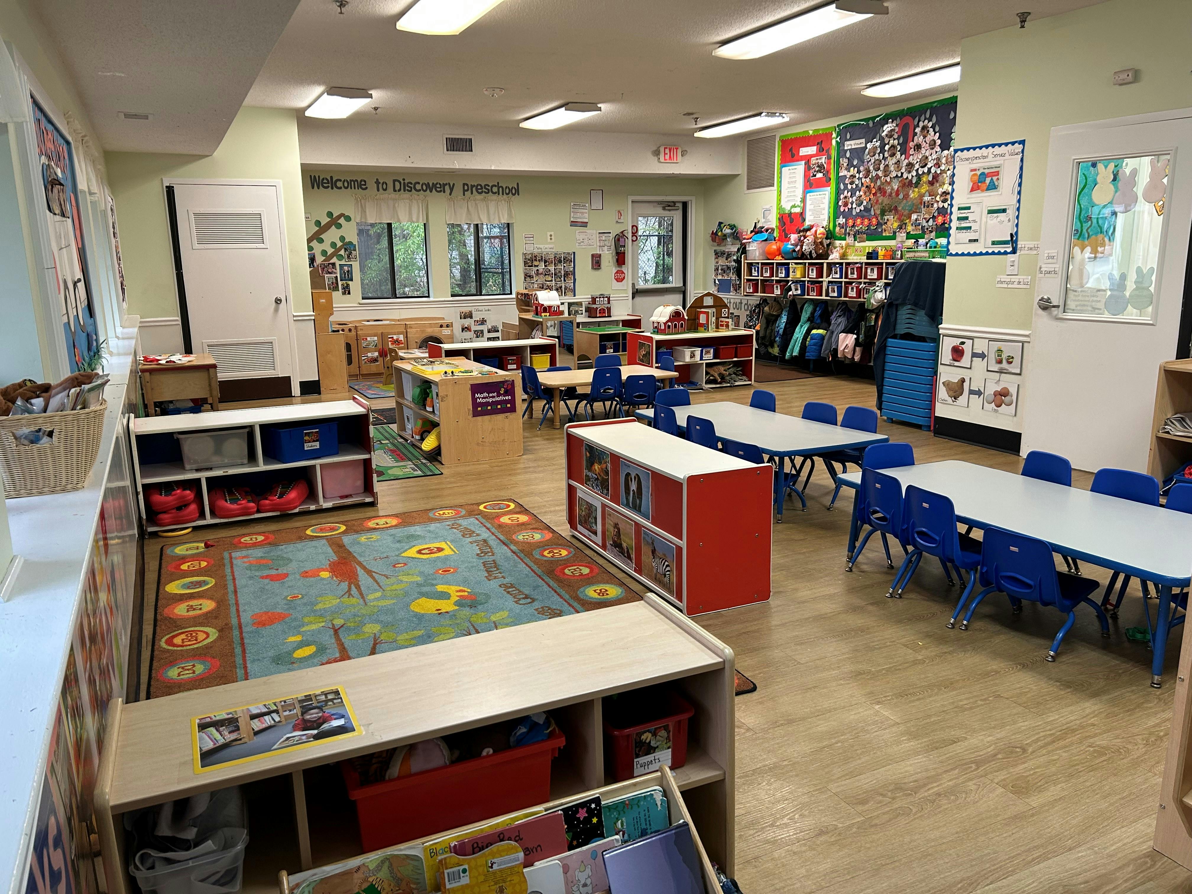An empty preschool classroom with small tables and chairs, a play kitchen, and colorful rugs