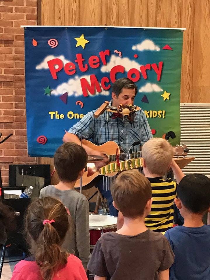 Person playing guitar and harmonica for children, "Peter McCory" banner in background