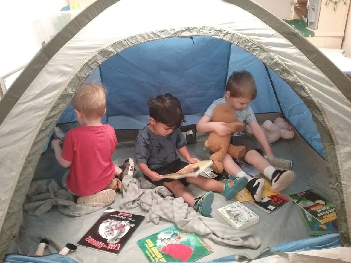 Three children sitting inside a tent with books and stuffed animals
