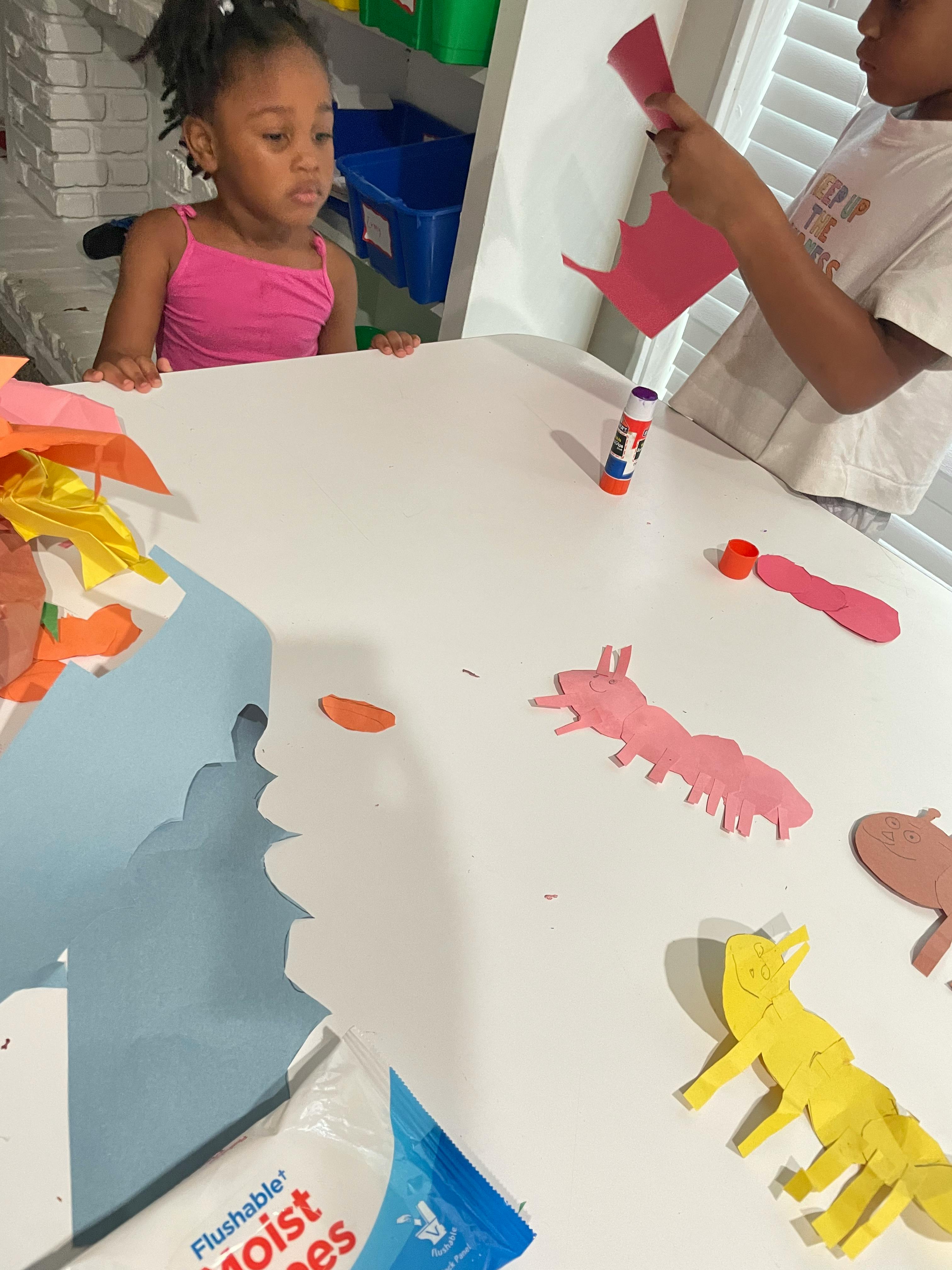 Child watching another child making paper cutouts at a table