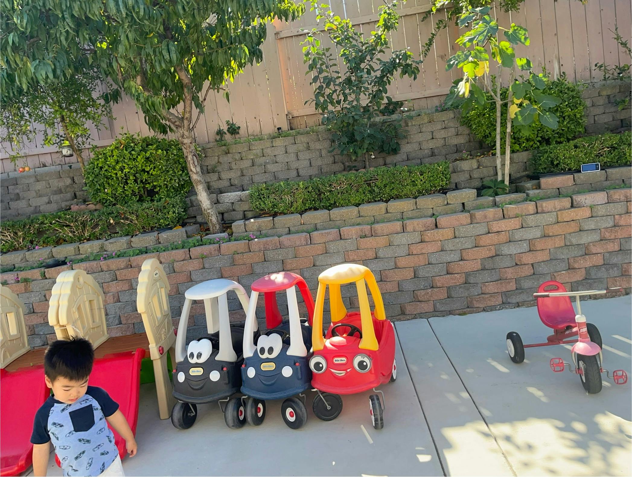 Child standing next to toy cars and a tricycle outdoors