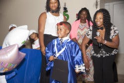 Child in graduation gown holding a microphone with adults standing adults
