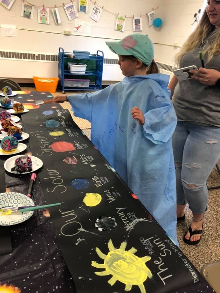 Child in a blue smock painting a solar system display