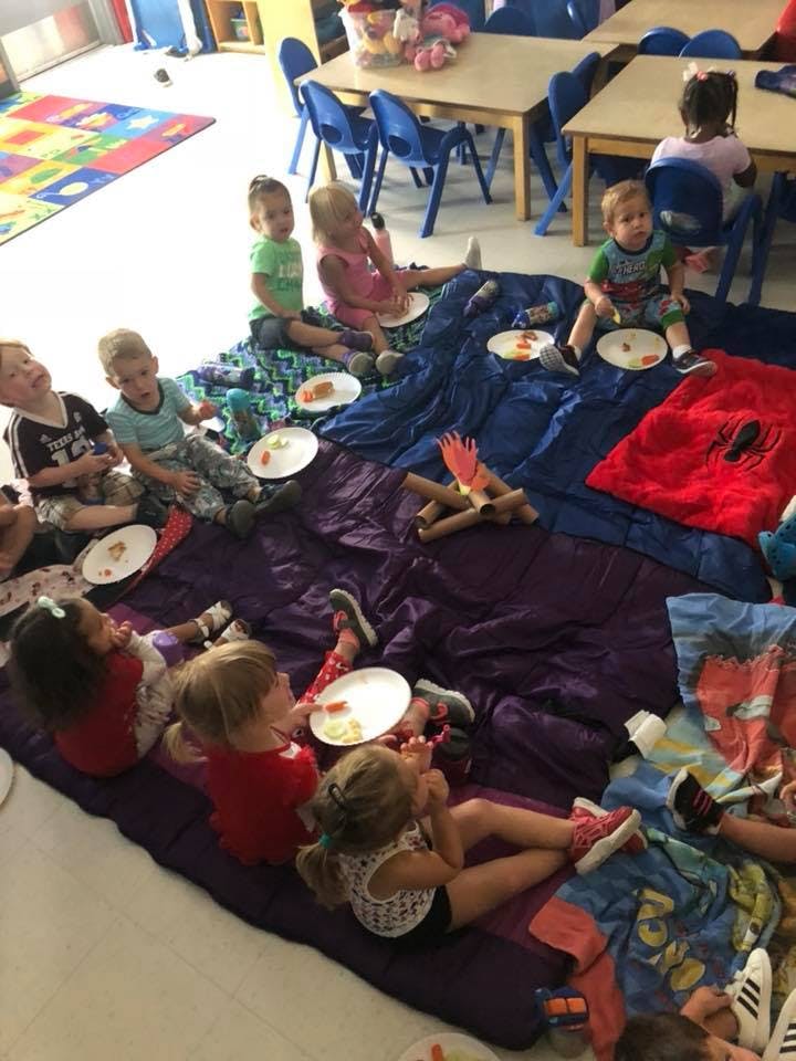 Children sitting on blankets with plates, some with a pretend campfire