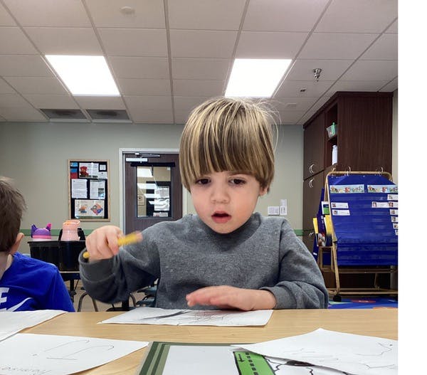 Child at a table holding a yellow pencil over a drawing