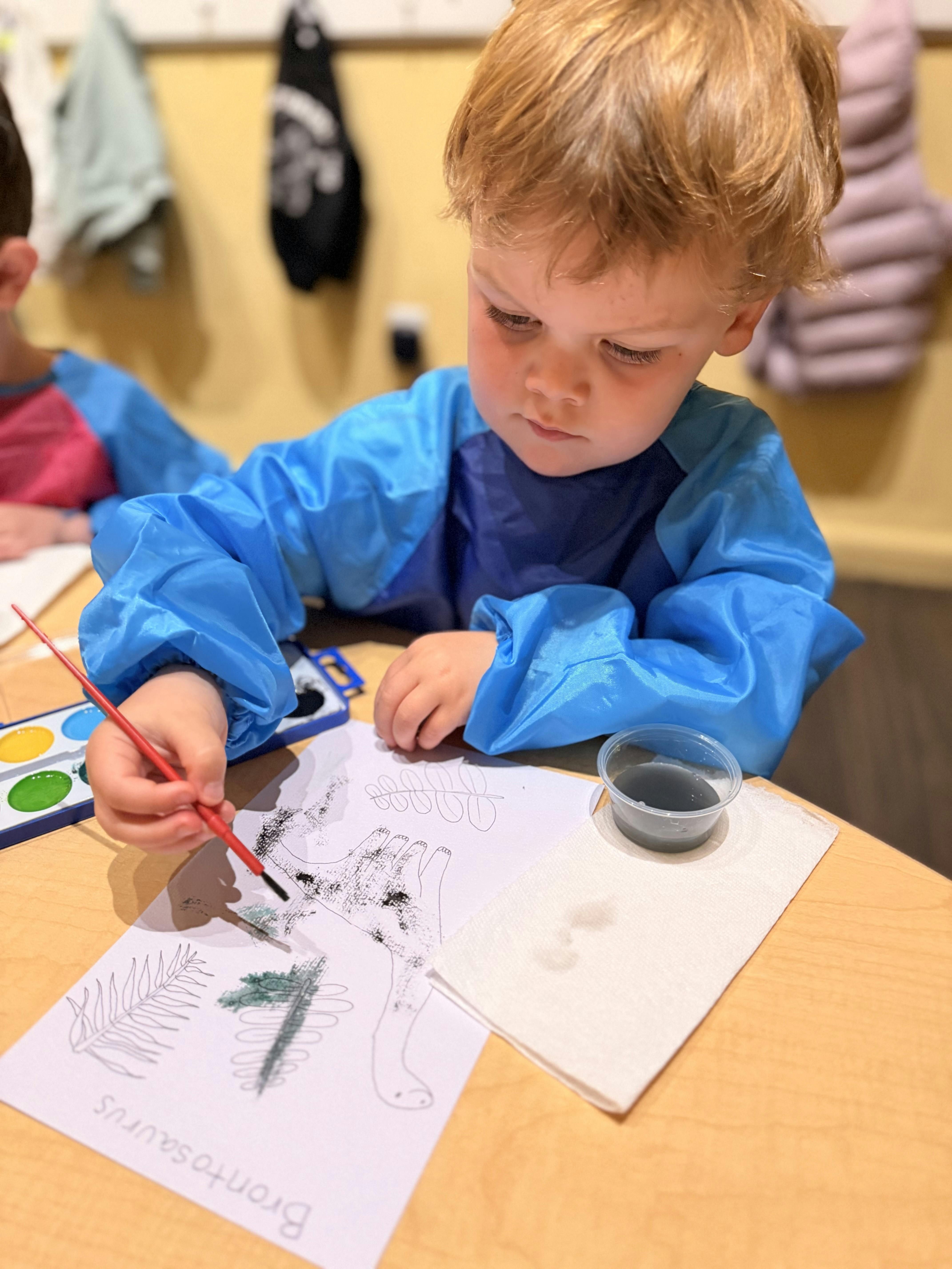 Child wearing an apron painting a dinosaur drawing at a table