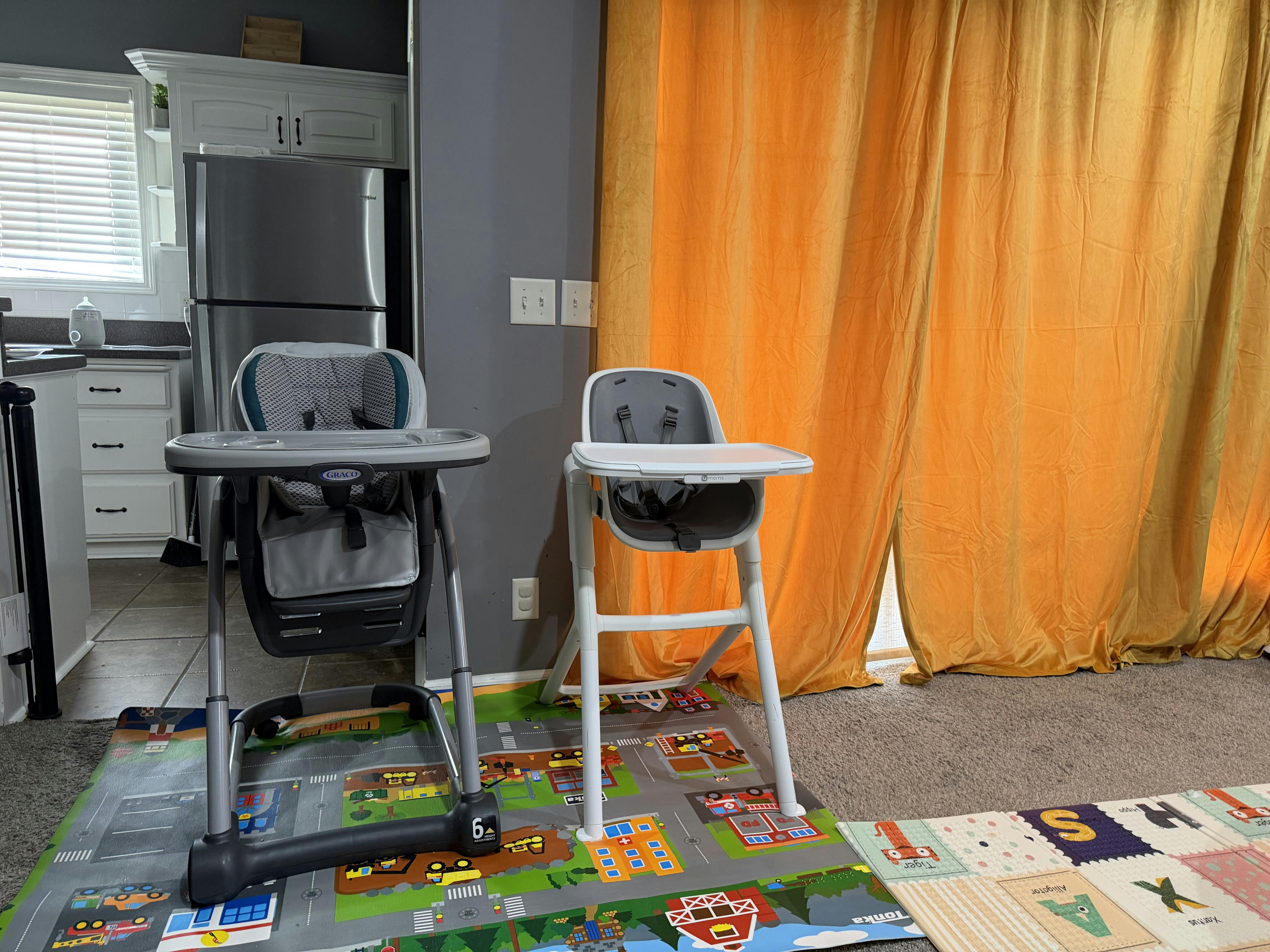 Two baby high chairs on playmats in a room with a kitchen in the background