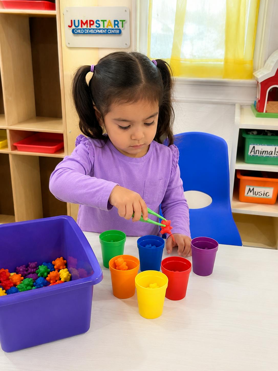 Child using tweezers to sort small colorful toys into cups in a classroom