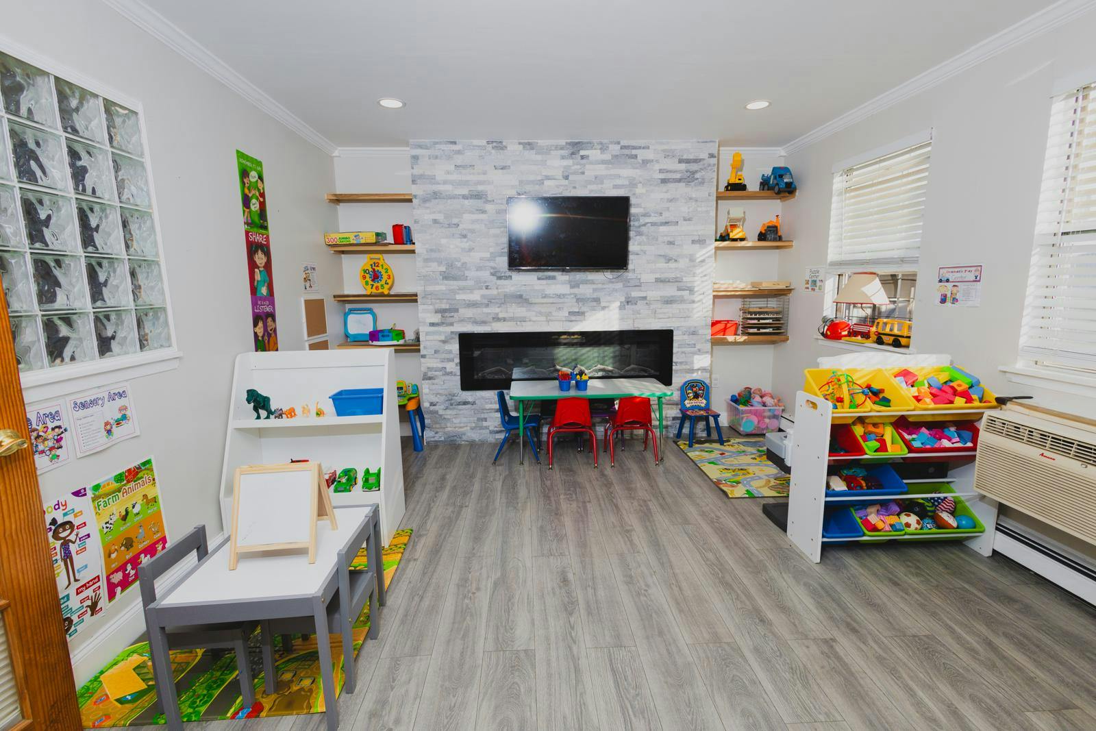 Playroom with small tables and chairs, shelves with toys, and a TV over a fireplace