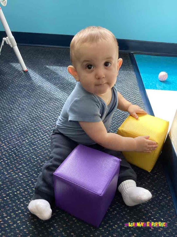 Child sitting on a purple block and holding a yellow block