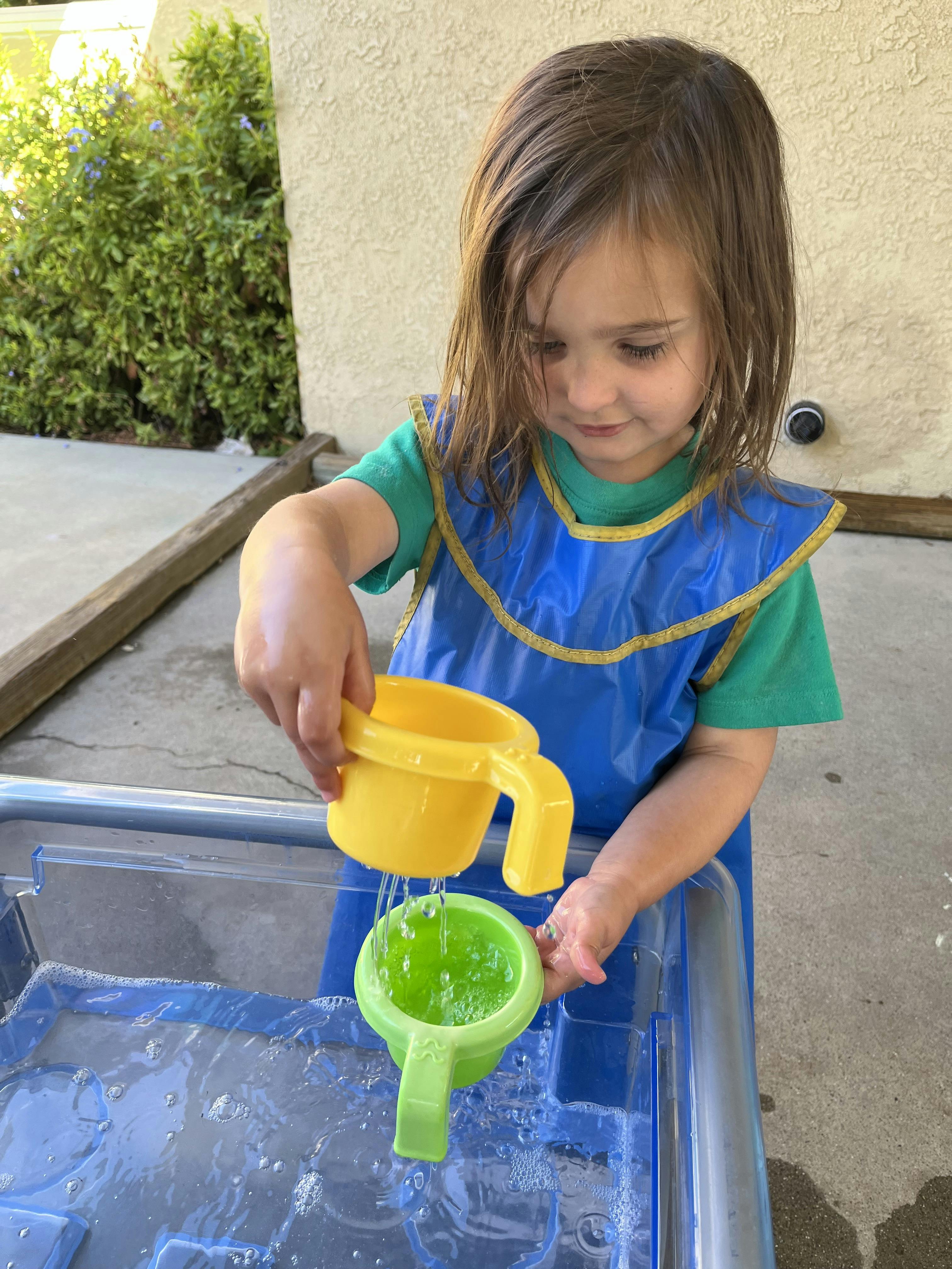 Child pouring water between two plastic cups over a water bin