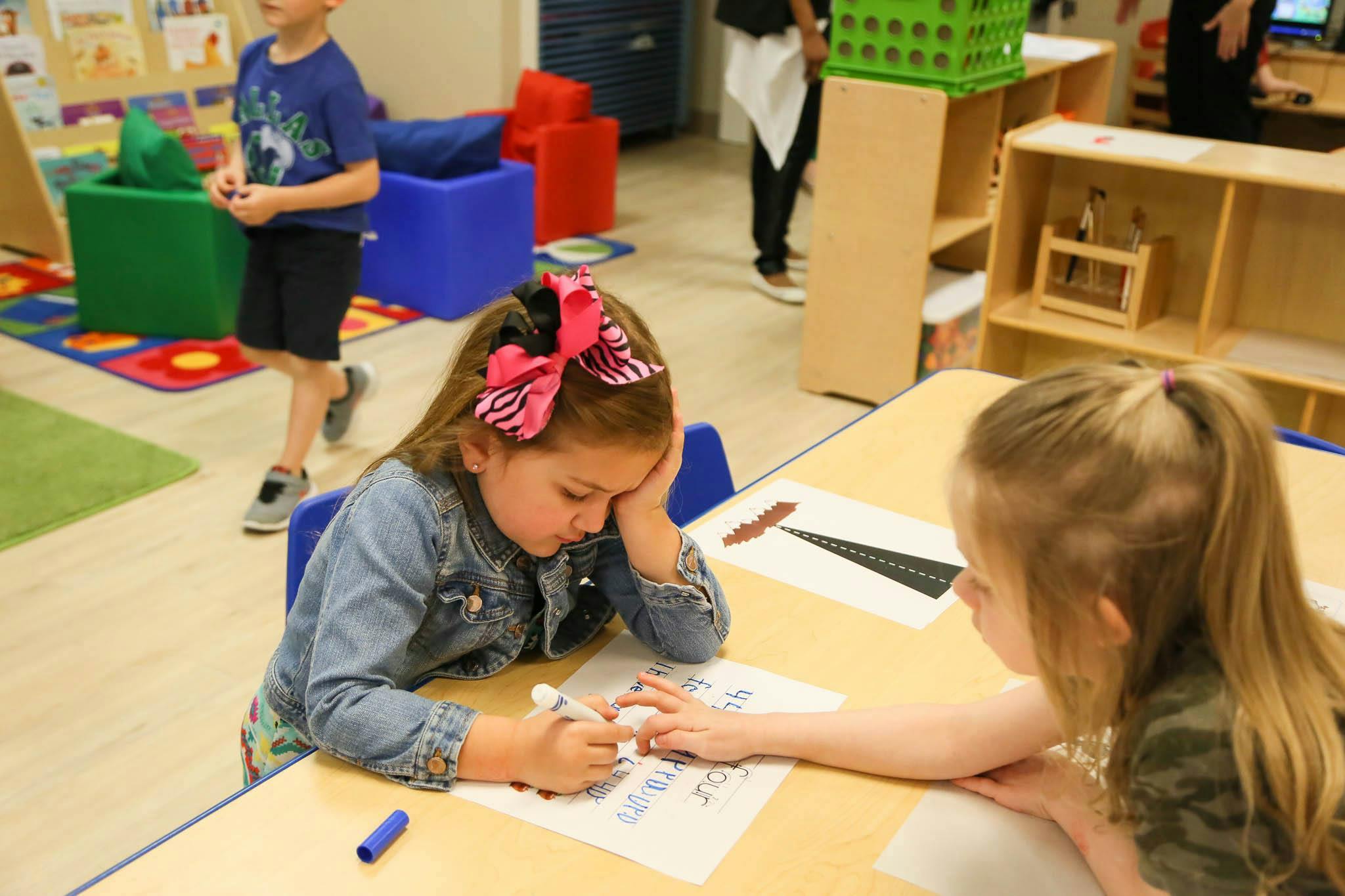 Child drawing on another child's hand at a table in a classroom