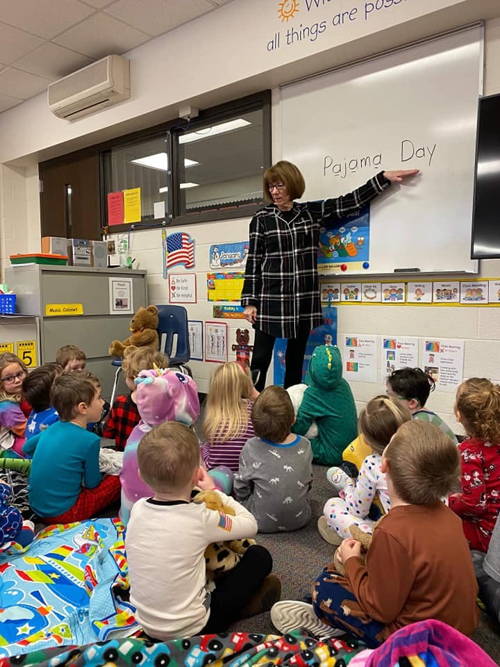 Person pointing at a whiteboard with "Pajama Day" written on it, surrounded by children
