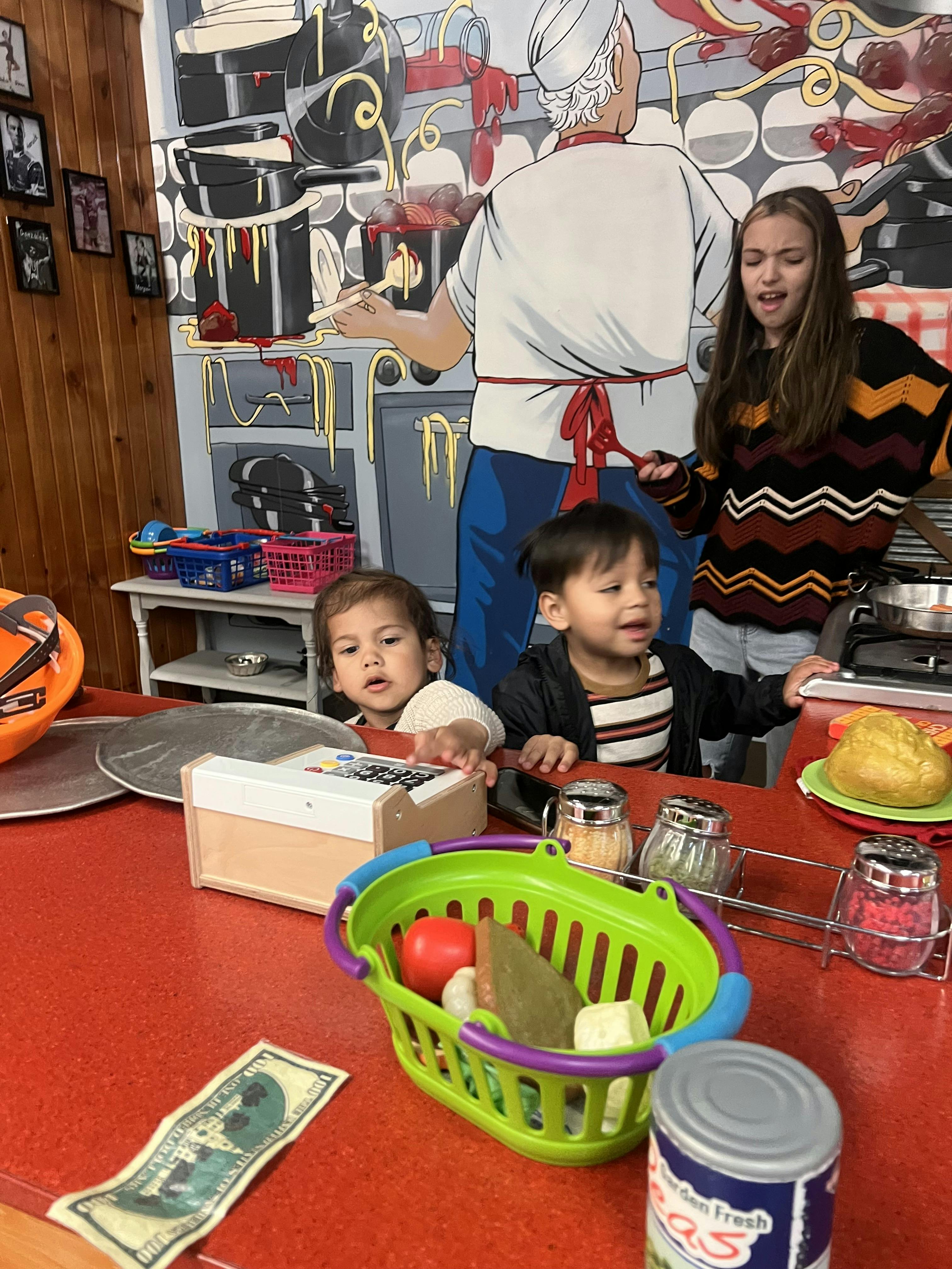 Two children and a teen playing with toy kitchen items and a cash register