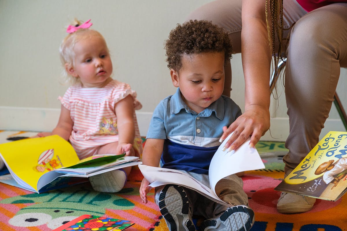 Two children sitting on a rug with books, one person turning a page