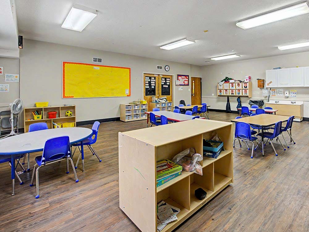 Classroom with multiple tables and blue chairs, shelves, and a yellow bulletin board
