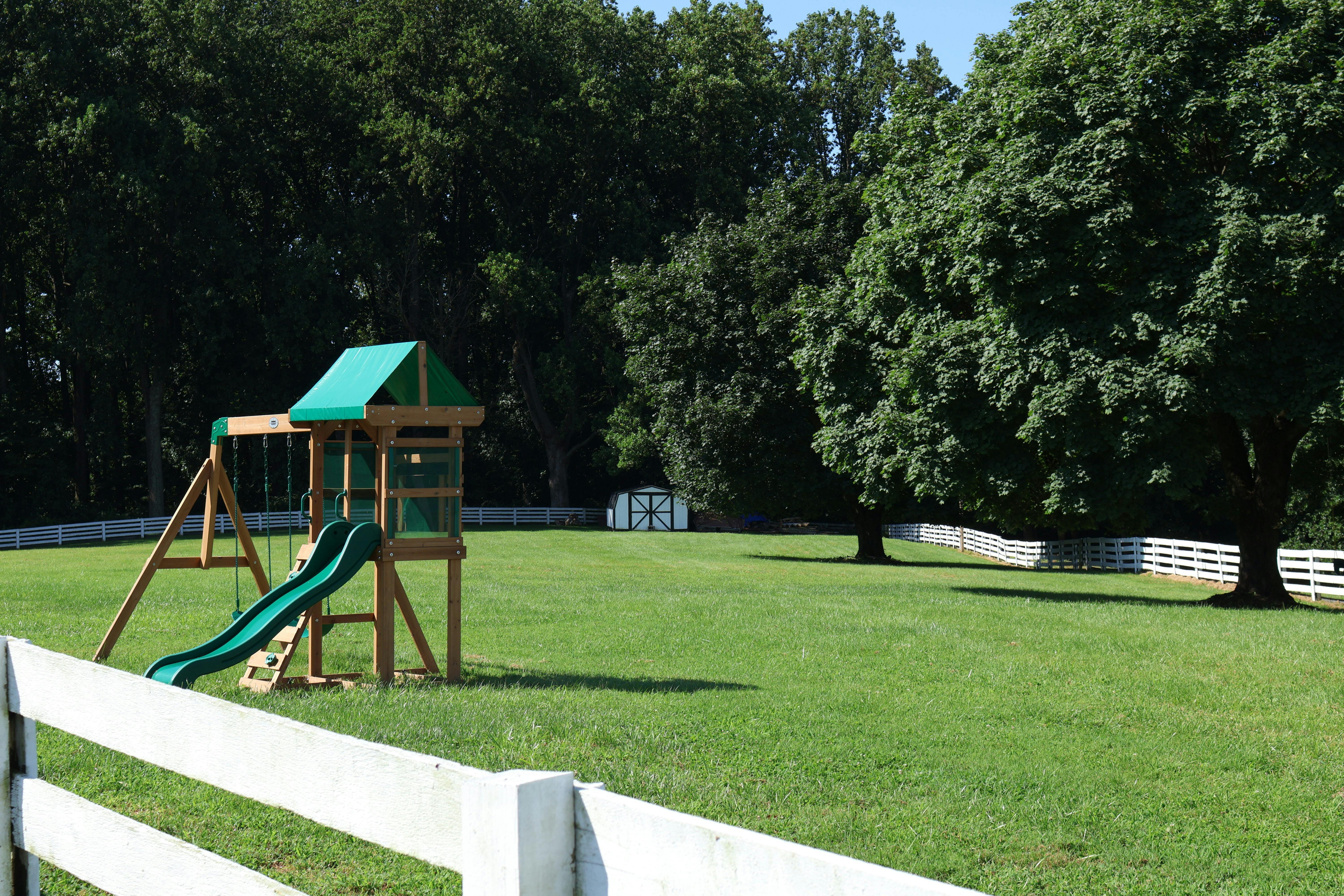 Playset with slide and swings in a grassy yard with trees and a white fence