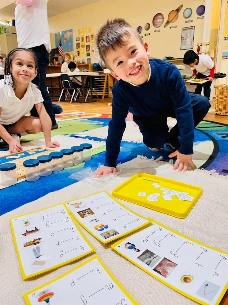 Two children on a colorful rug in a classroom, one smiling at the camera