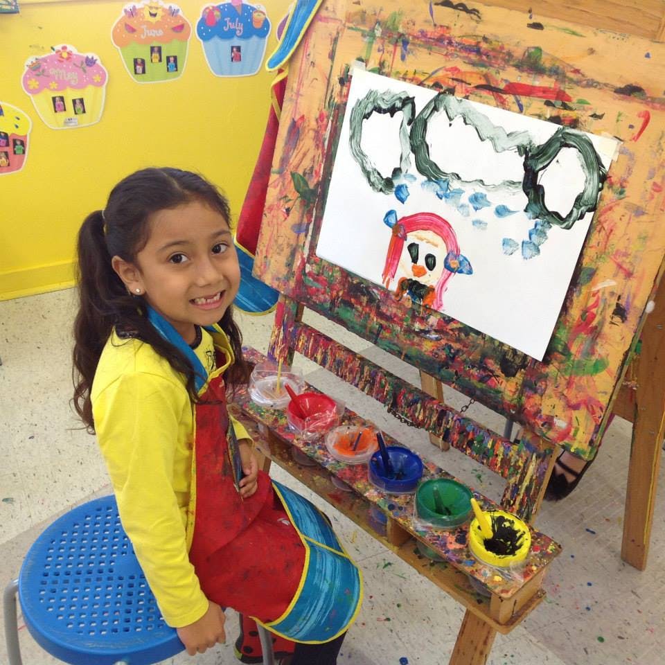 Child in an apron sitting next to an easel with a painting