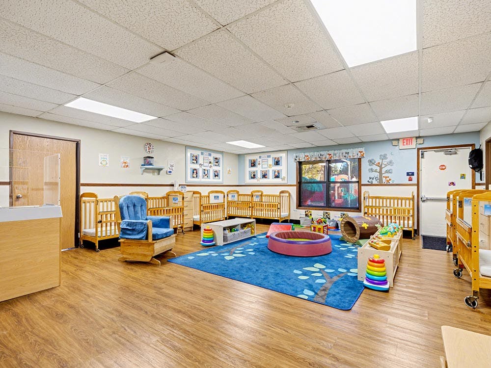 A brightly lit nursery with cribs, a blue rug, and various toys