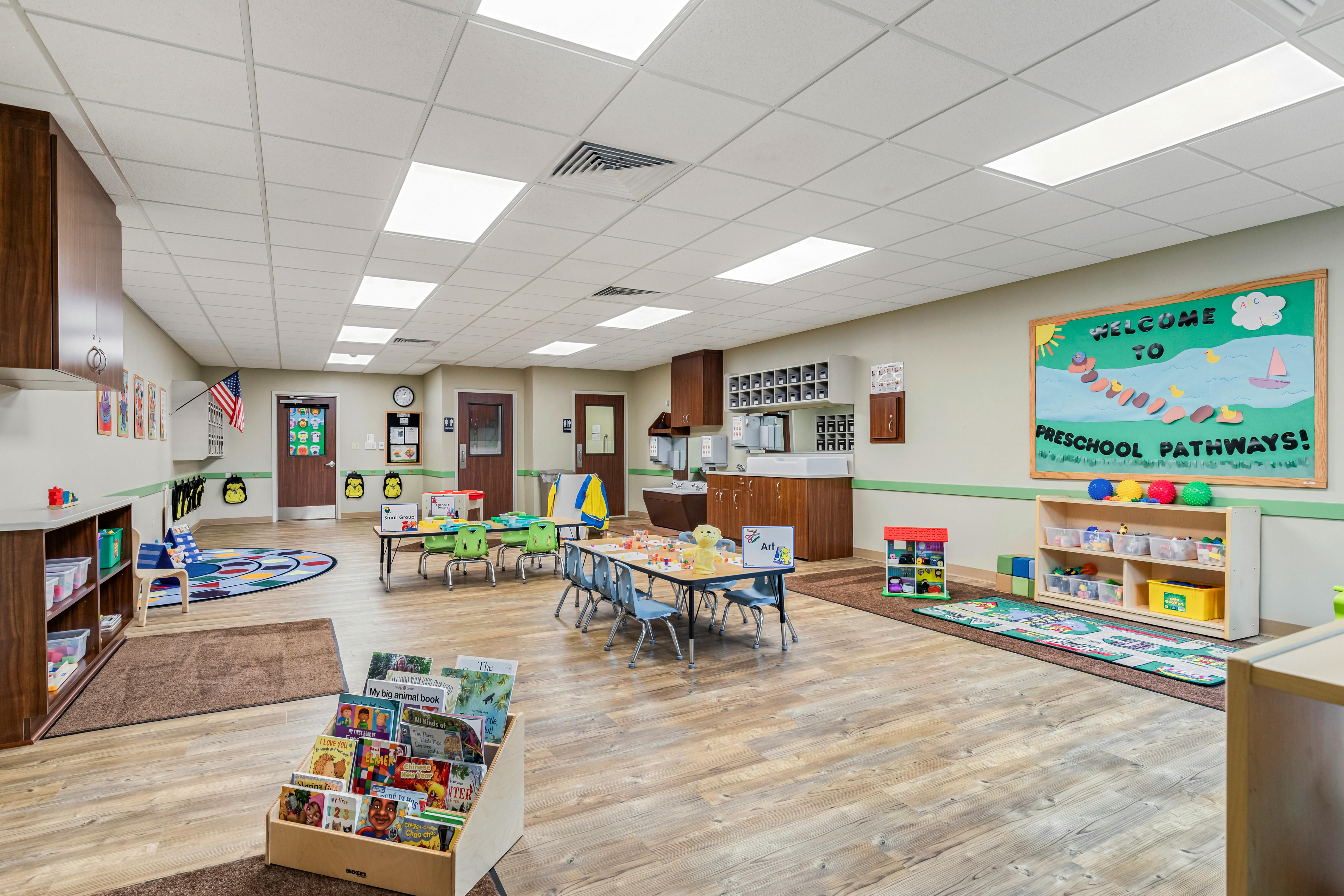 An empty preschool classroom with small tables and chairs, a play rug, and shelves