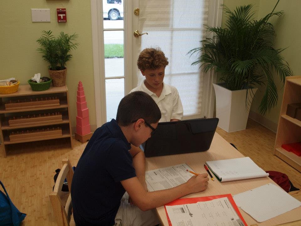 Two children working at a table, one writing and one looking at a laptop
