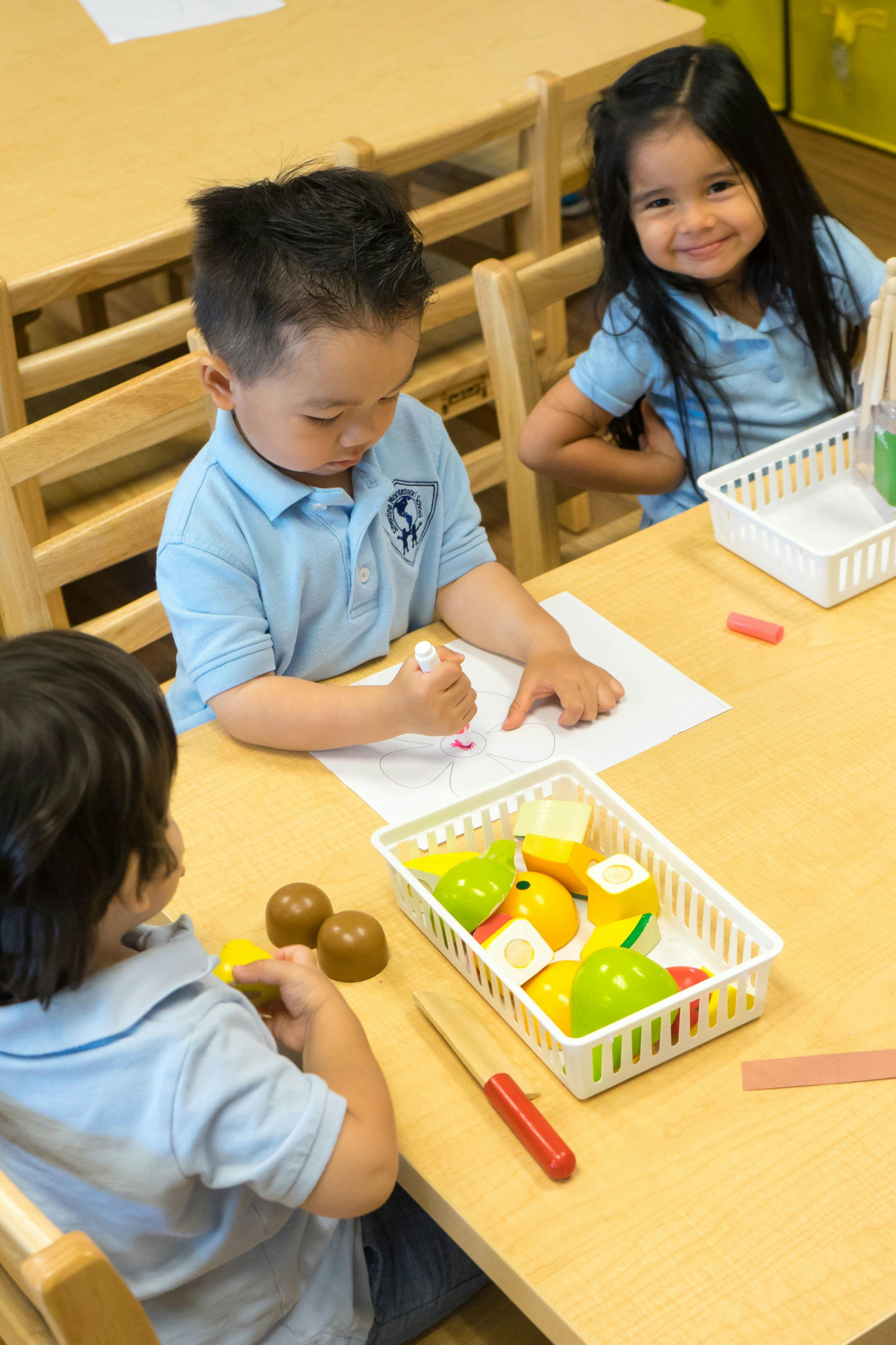 Three children at a table, one drawing, one playing with toy food