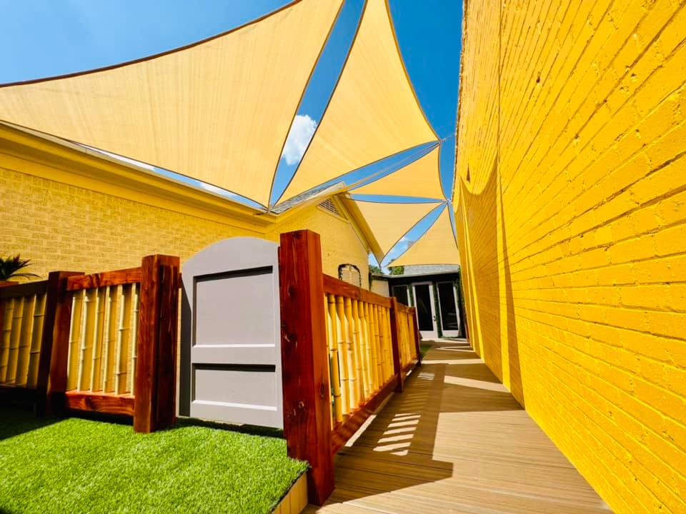 Outdoor area with yellow shade sails, a wooden fence, and artificial grass
