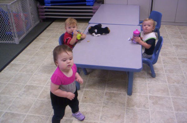 Three children in a classroom, two seated at a table with bottles