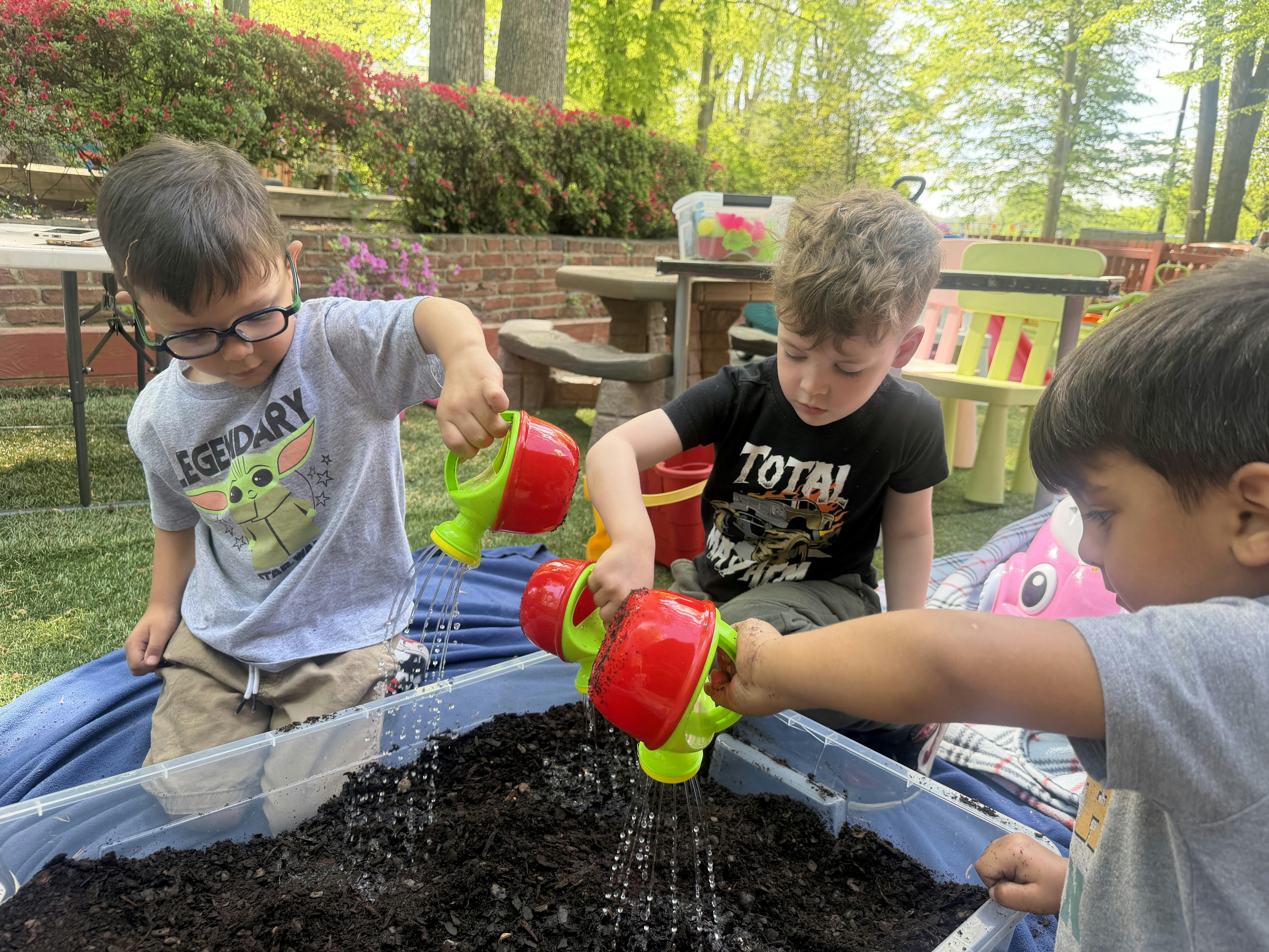 Three children watering dirt in a plastic bin with toy watering cans