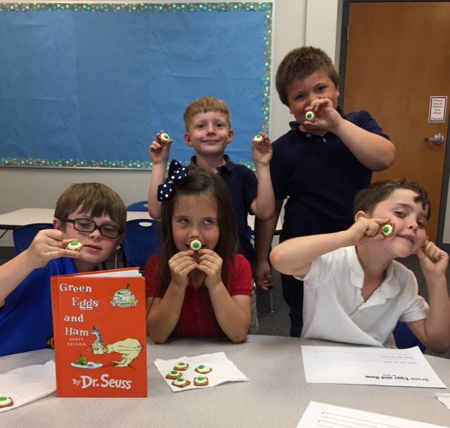 Children holding up cookies with green frosting, one child holds a book
