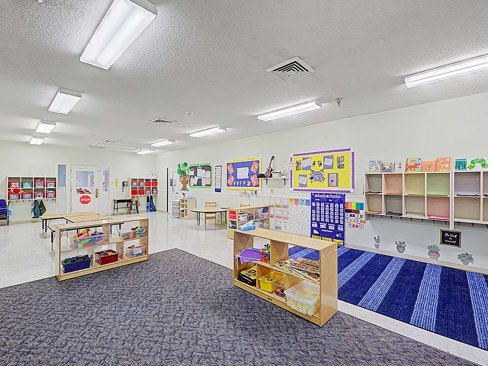 A brightly lit classroom with tables, cubbies, shelves of toys, and wall displays