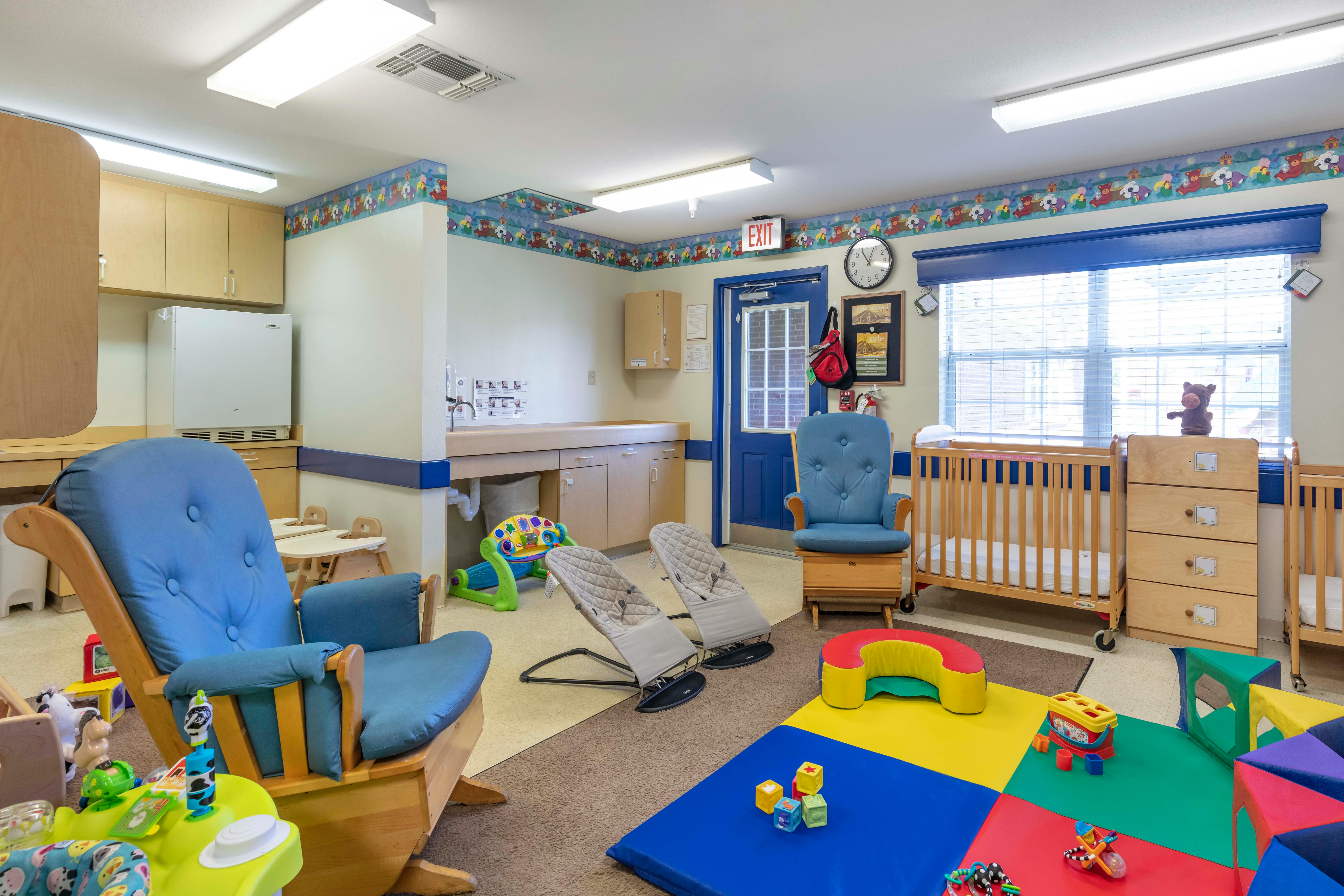 A brightly lit daycare or nursery room with cribs, rocking chairs, and play mats