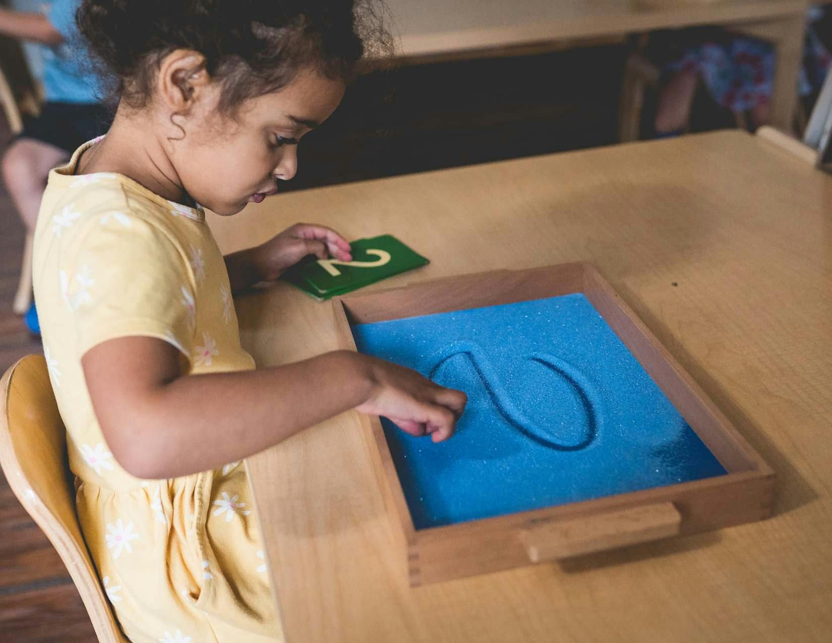 Child tracing a letter in a sand tray with a number card nearby