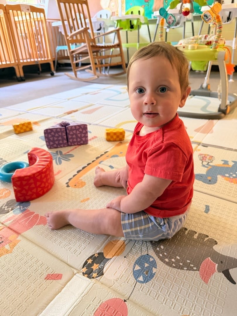 A toddler in a red shirt sitting on a play mat with toys