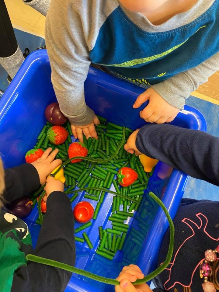 Children playing with toy vegetables and green pipe cleaners in a blue bin
