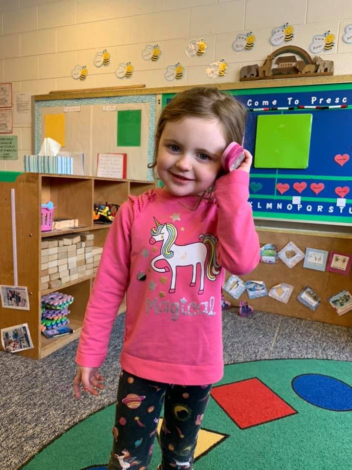 Child in a classroom holding a pink object to their ear